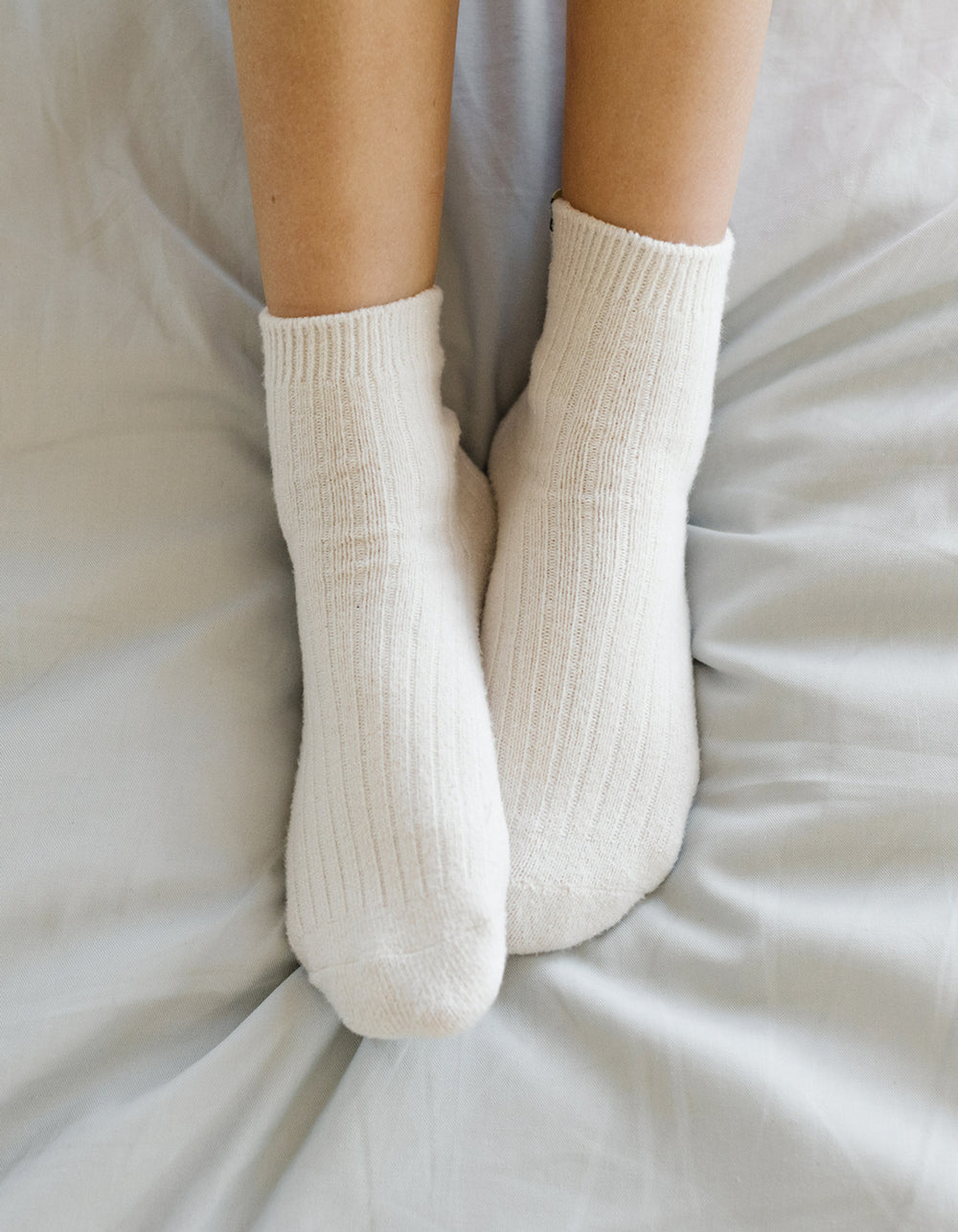 A close-up of two feet wearing Cozy Earth's Plush Modern Crew Sock 3-Pack in white, resting on a light gray bedsheet.
