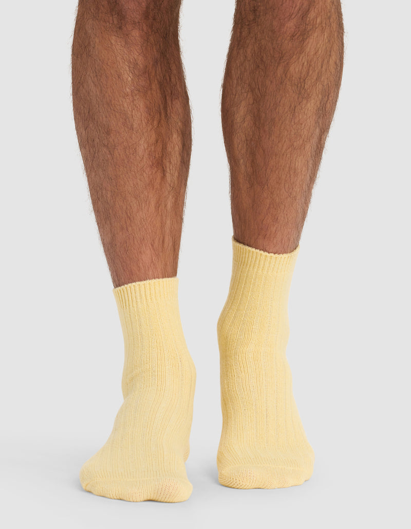 A person stands on a light surface, shown from the calves down, wearing Cozy Earth's Plush Lounge Quarter Socks in pale yellow. Their feet are close together, set against a neutral gray background.