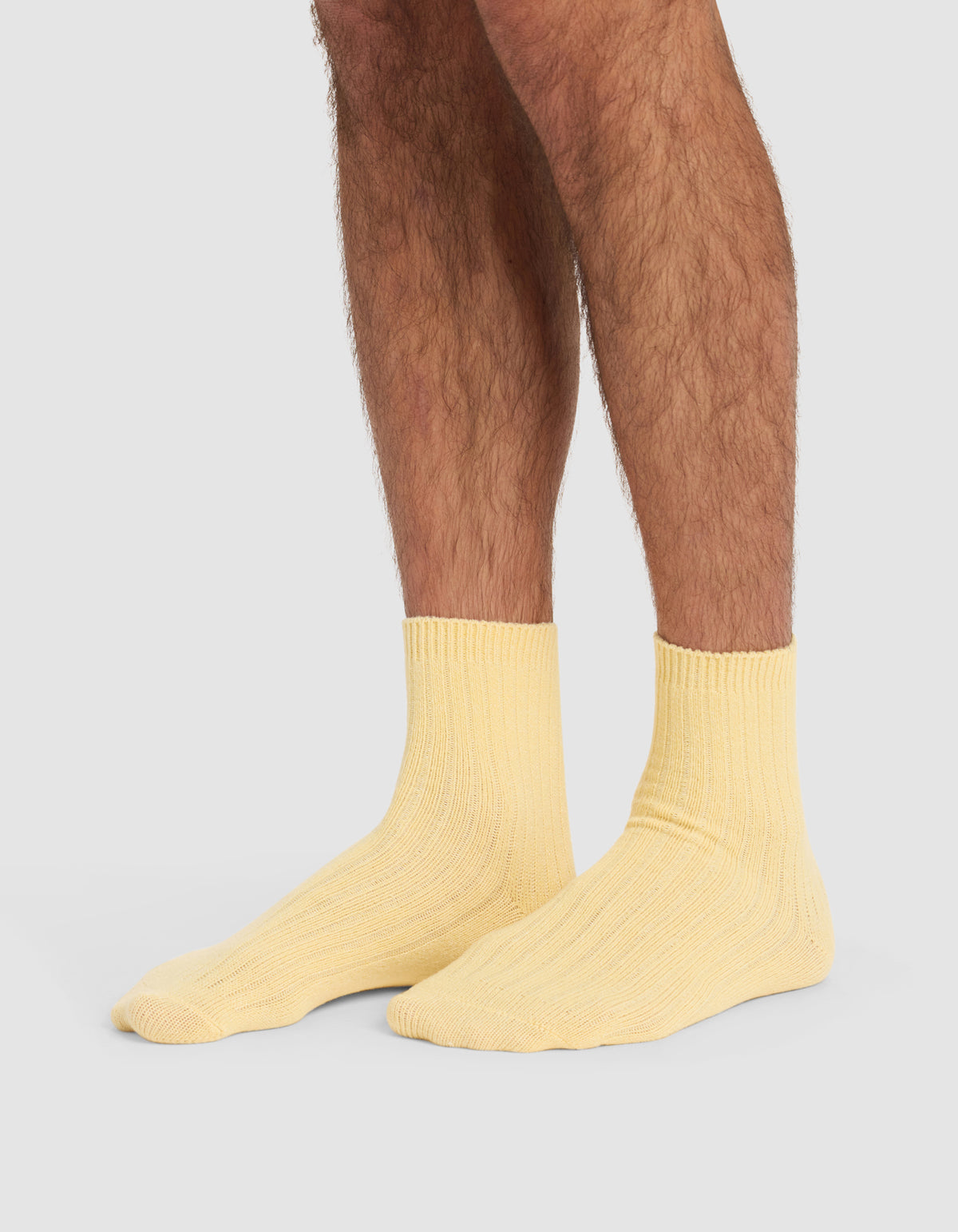 A person with hairy legs is wearing Cozy Earth's The Plush Modern Crew Sock in light yellow, standing on a plain, light grey background.