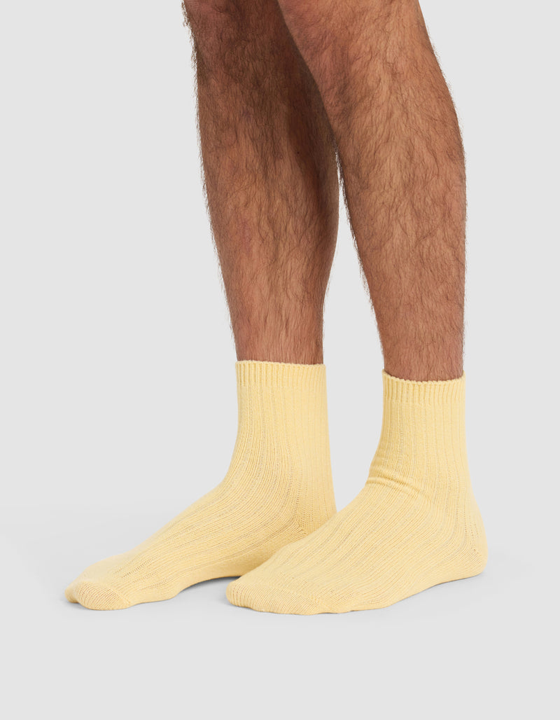 A person with hairy legs is wearing Cozy Earth's Plush Lounge Quarter Socks in light yellow, standing on a plain, light grey background.