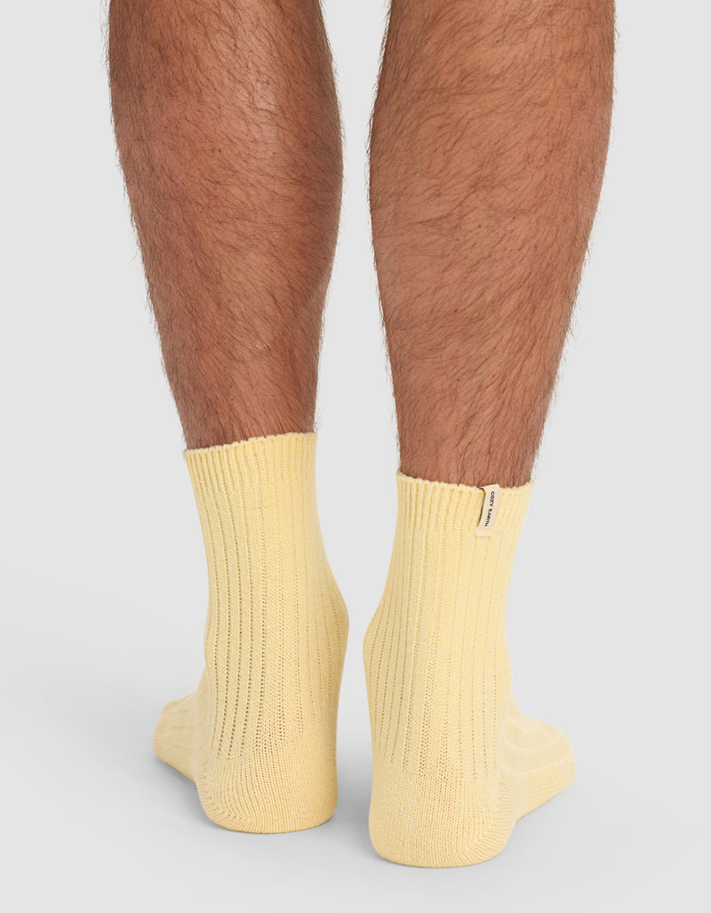 The back view of a person's lower legs wearing Cozy Earth's Plush Lounge Quarter Socks in pale yellow ribbed fabric, standing on a white surface.
