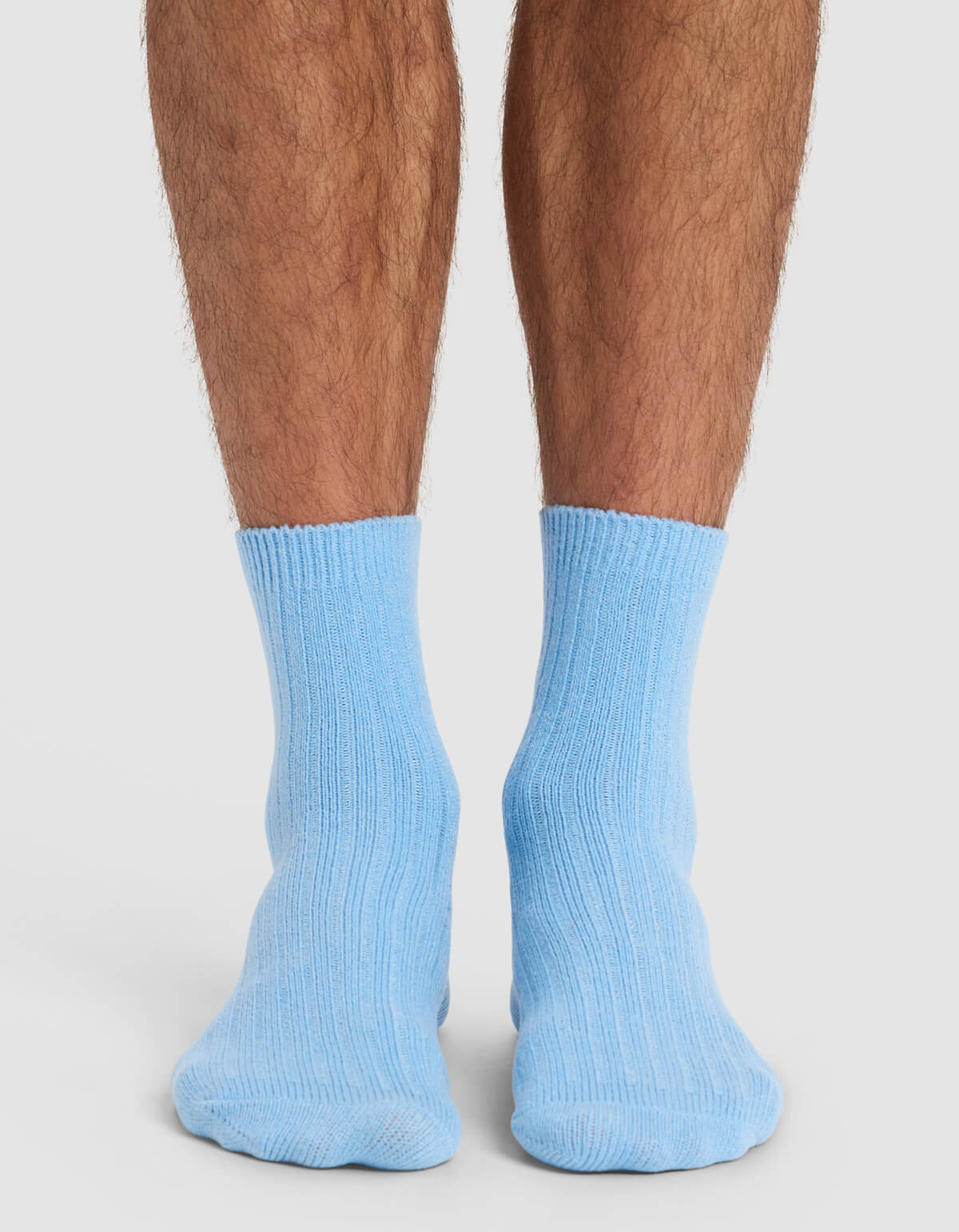 A close-up of a person's lower legs and feet, seen from behind, wearing Cozy Earth's Men's Plush Lounge Crew Sock in light blue. The socks are ribbed and the person is standing on a white surface.