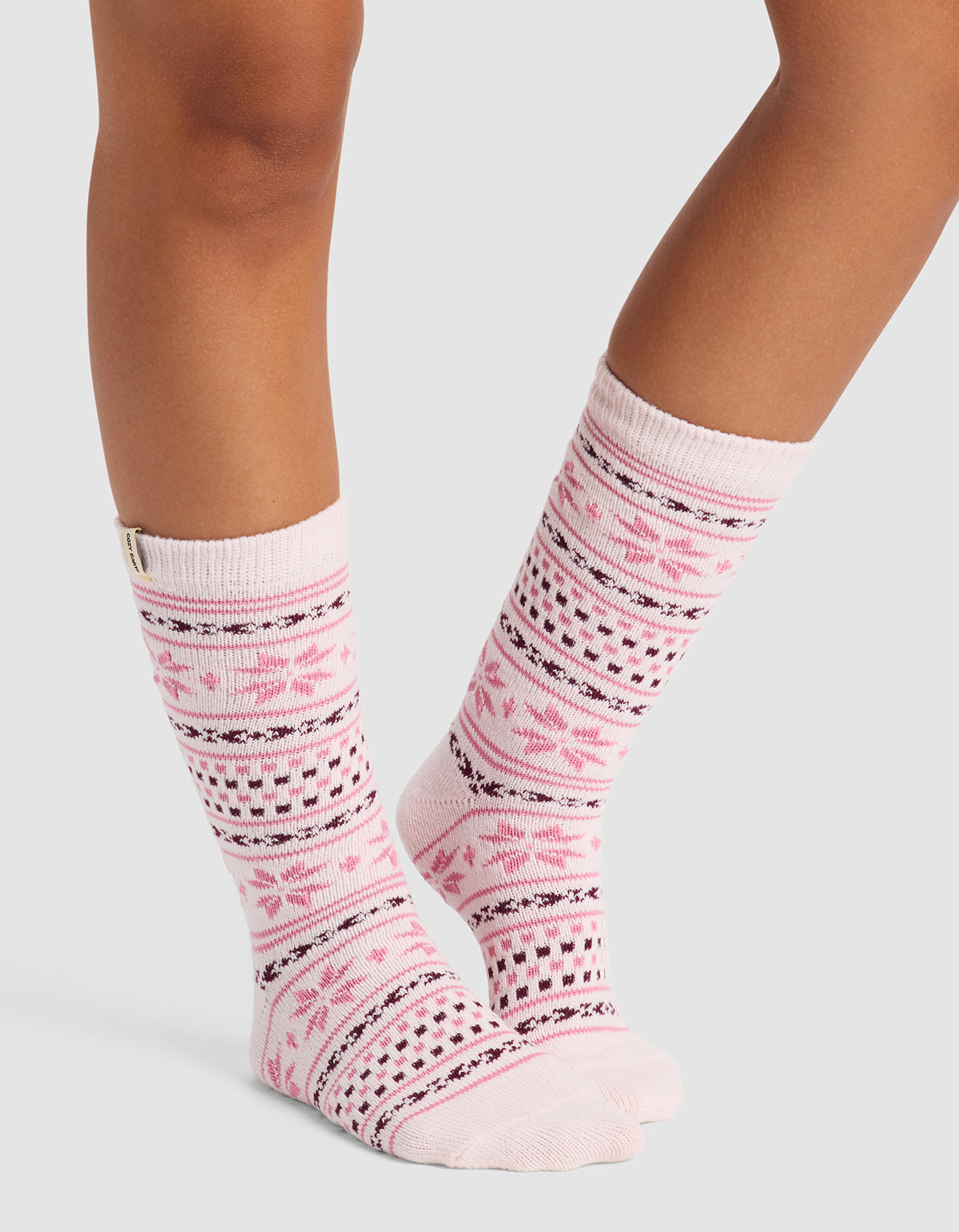 A person stands on a light gray background wearing Cozy Earth's Snowflake Plush Lounge Sock, a knee-high pink and white sock with Nordic snowflakes and geometric patterns.