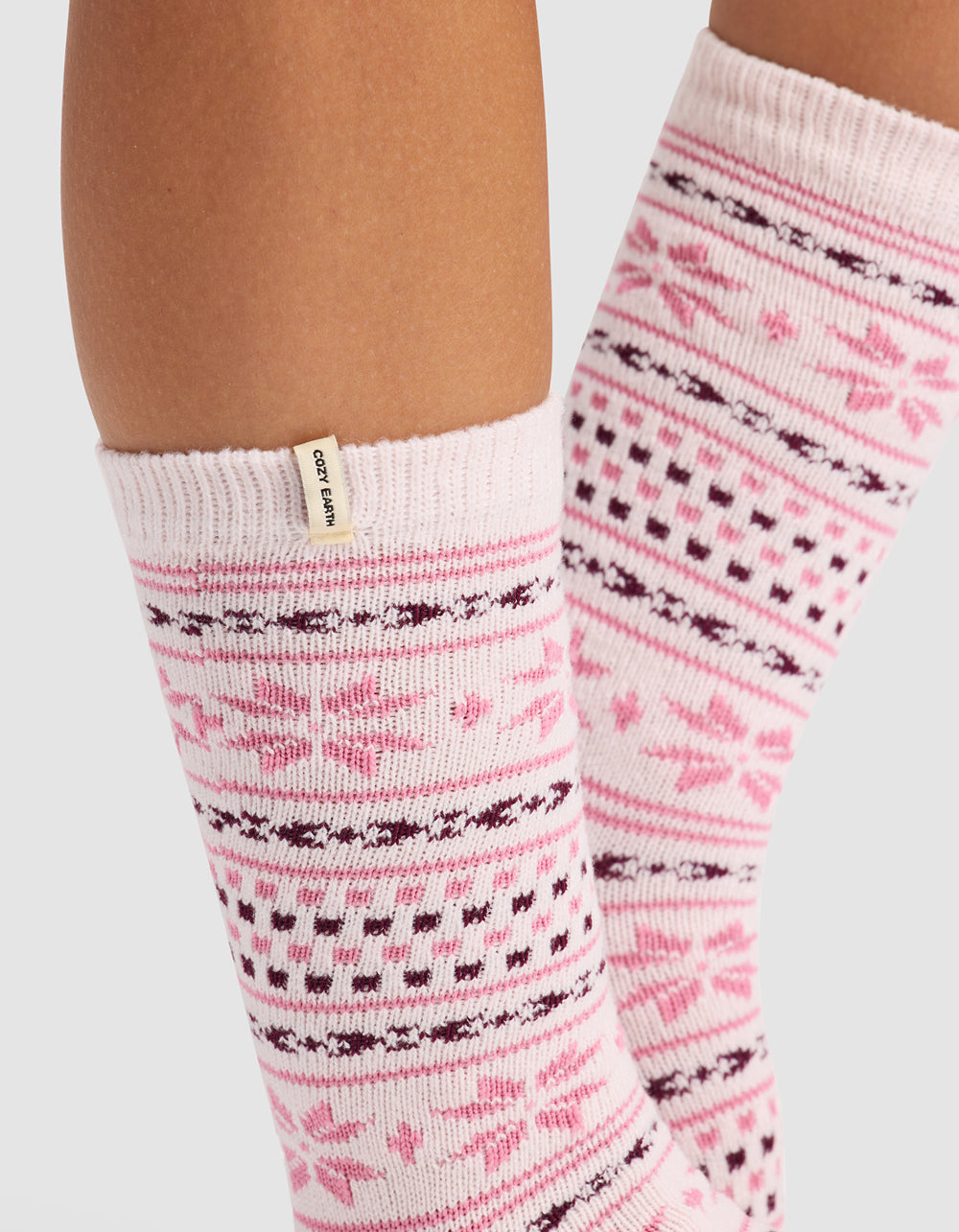 Close-up of a person's lower legs wearing Cozy Earth's The Snowflake Plush Lounge Sock in light pink with dark pink and burgundy snowflake and geometric patterns, featuring a small beige tag on the sock cuff.