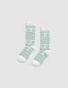 The Snowflake Plush Lounge Sock by Cozy Earth, a pair of white crew socks with green and blue geometric and floral patterns, displayed flat on a plain light gray background.|Color:Celadon