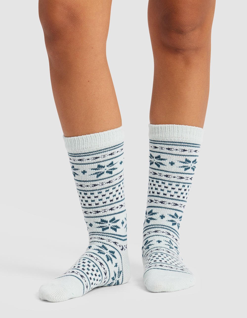 A person stands on a plain white background wearing The Snowflake Plush Lounge Sock by Cozy Earth—knee-high, white socks featuring blue geometric and snowflake patterns.