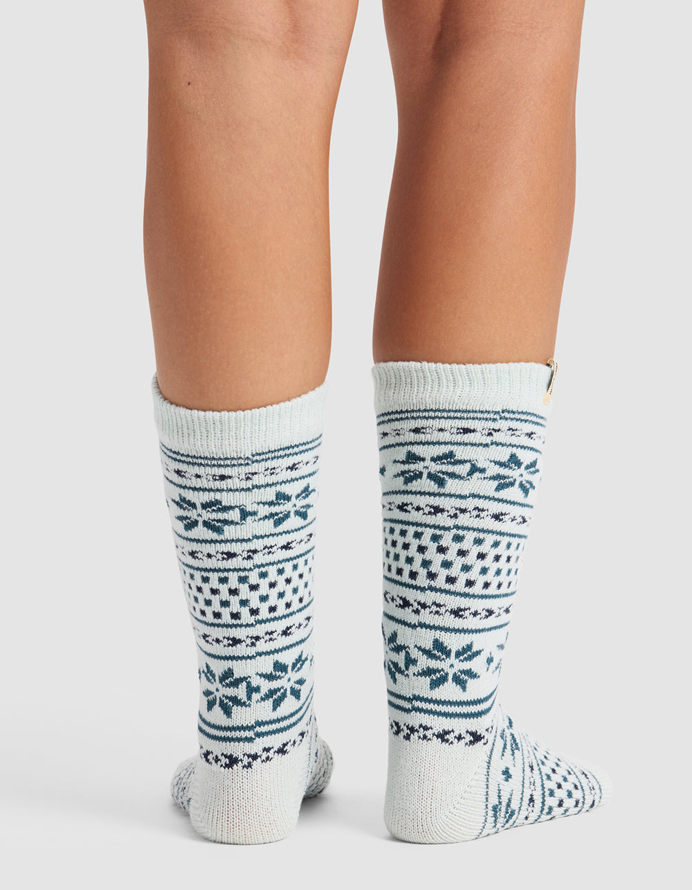 A person wears The Snowflake Plush Lounge Sock by Cozy Earth—knee-high, white knit socks with blue and gray geometric and snowflake patterns—standing with feet together against a plain background.