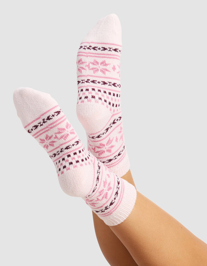 A person wears The Snowflake Plush Modern Crew Sock by Cozy Earth—light pink socks with darker pink and burgundy geometric and floral designs—feet raised against a plain, light gray background.