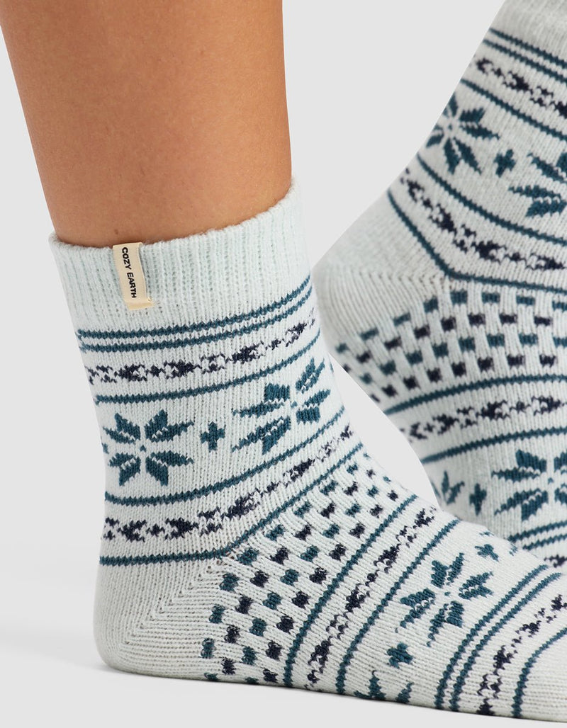A close-up of feet in The Snowflake Plush Modern Crew Sock by Cozy Earth, featuring light-colored wool with blue geometric and snowflake patterns and a beige cuff tag labeled "Cozy Earth.