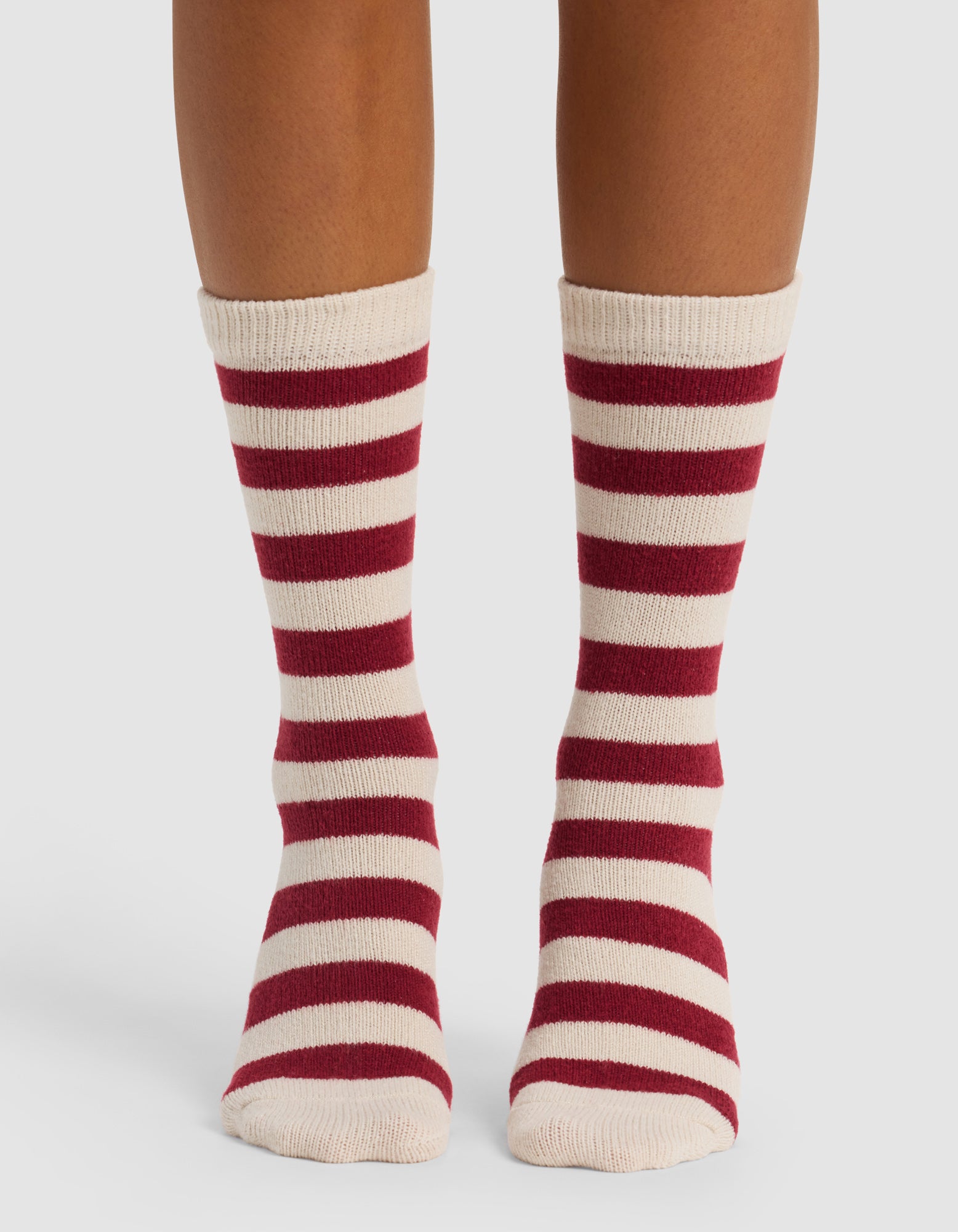 A person wearing Cozy Earth's The Striped Plush Lounge Sock in cream with red horizontal stripes stands on a white surface, showing only their lower legs and feet.|Color:Garnet Stripe