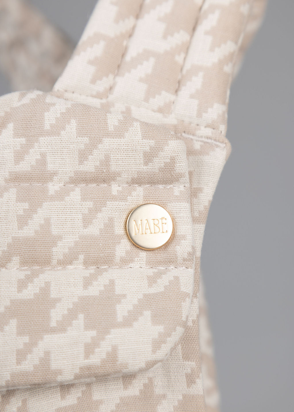 Close-up of The Monarch Carrier - Houndstooth by Mabē, showcasing beige and white houndstooth fabric with quilting and a gold button engraved with "Mabē.