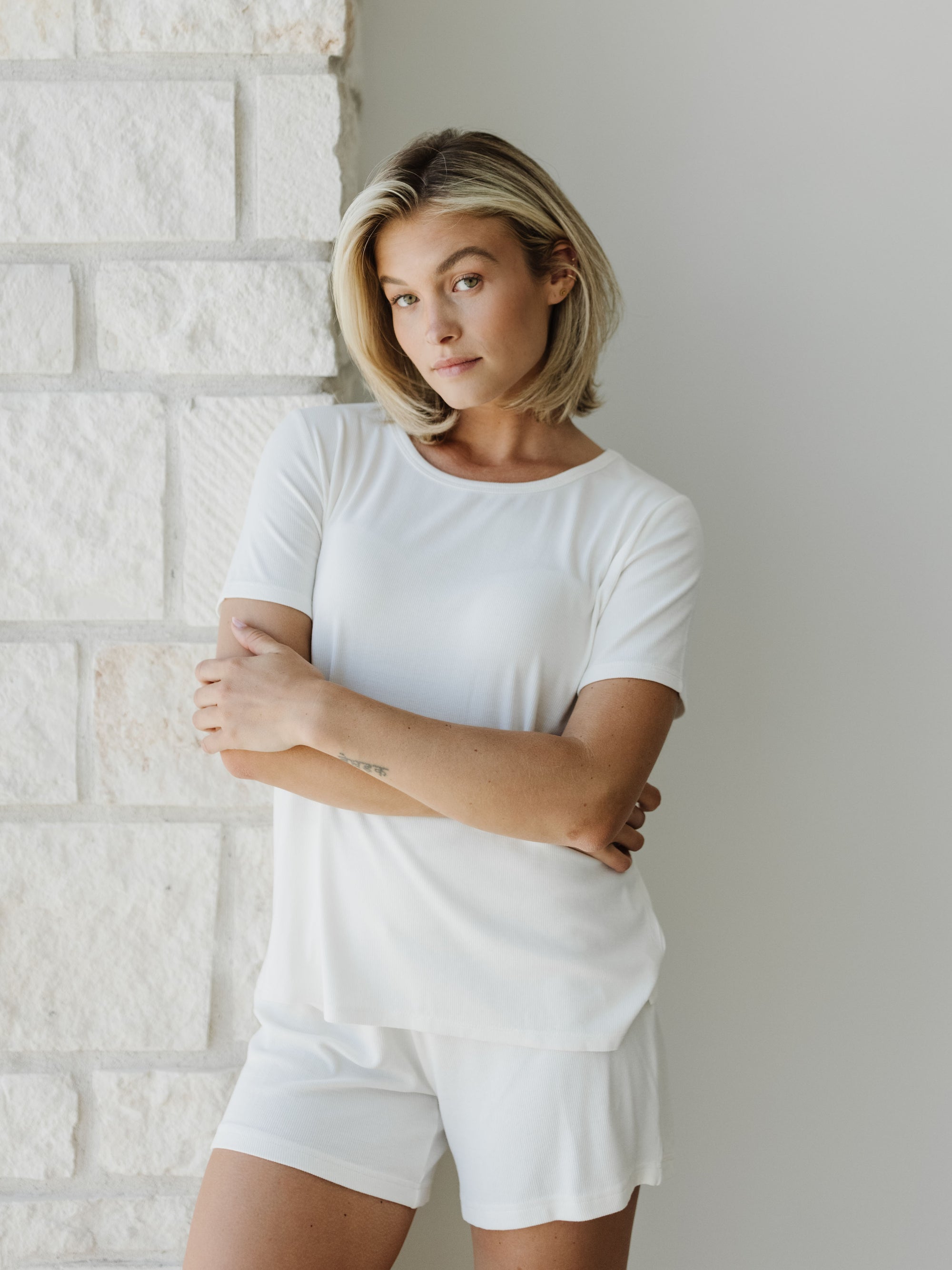 Woman wearing a white Cozy Earth Rib Knit Short Sleeve Lounge Top and white shorts against a white stone background 