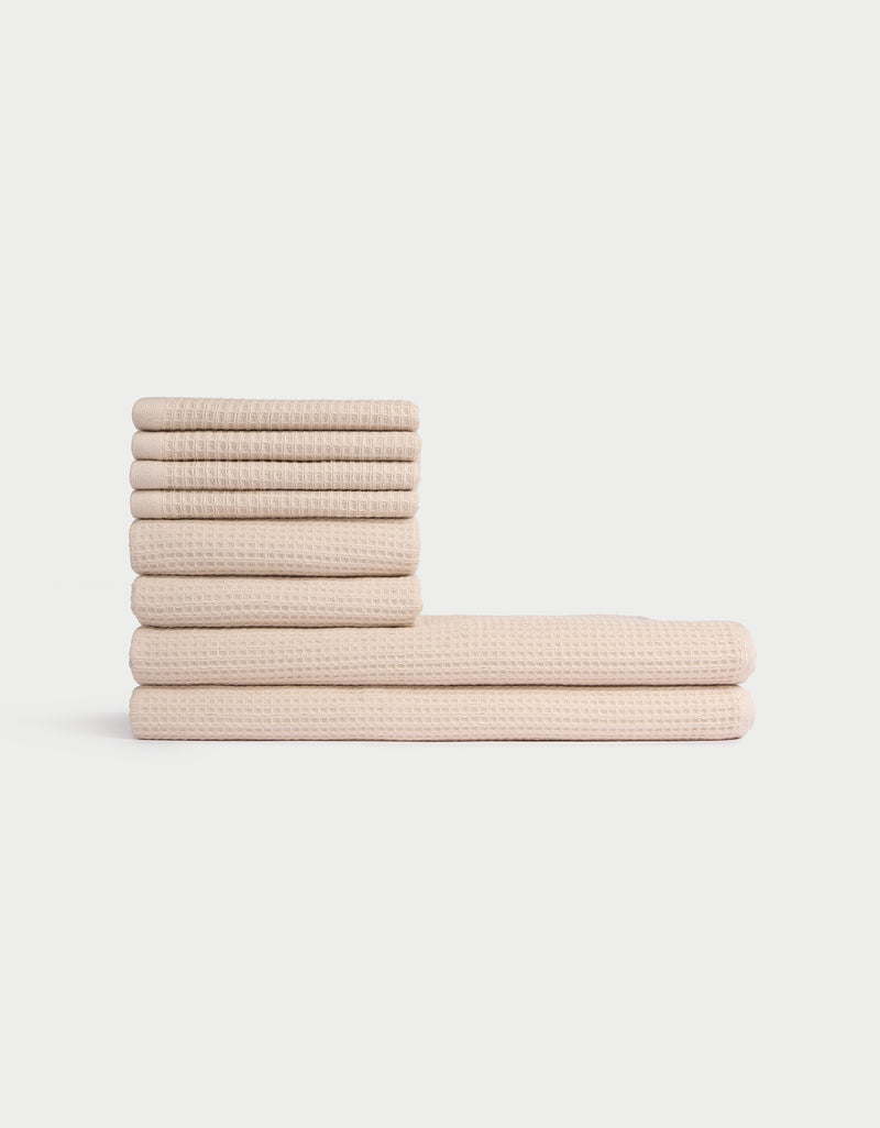 The Cozy Earth Waffle Bath Towel Set features eight neatly folded beige towels with a textured waffle pattern, stacked in two piles against a plain white background.