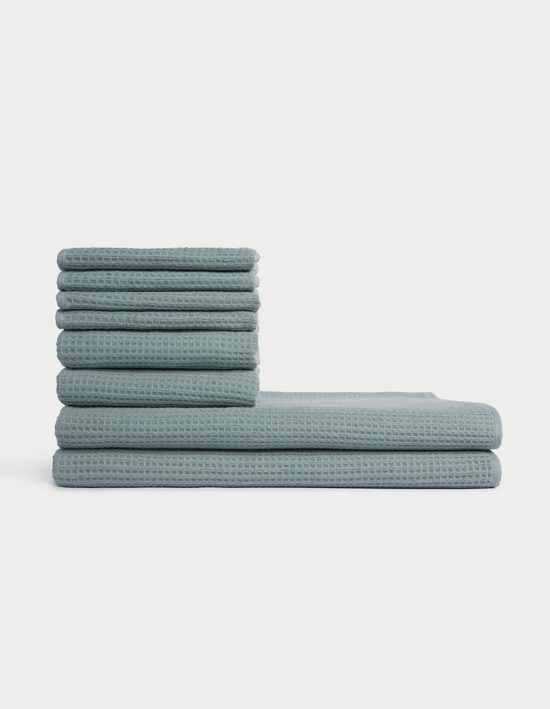 The Cozy Earth Waffle Bath Towel Set features blue, waffle-textured towels stacked in two vertical piles of varying heights against a plain white background.