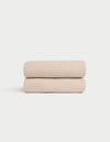 Two Cozy Earth Waffle Hand Towels in light beige are neatly folded and stacked against a plain white background, showcasing their textured pattern. |Color:Birch