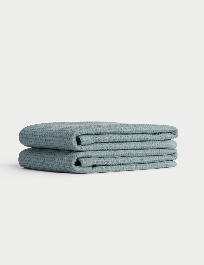 A neatly folded stack of two light blue Cozy Earth Waffle Hand Towels is displayed on a white background.