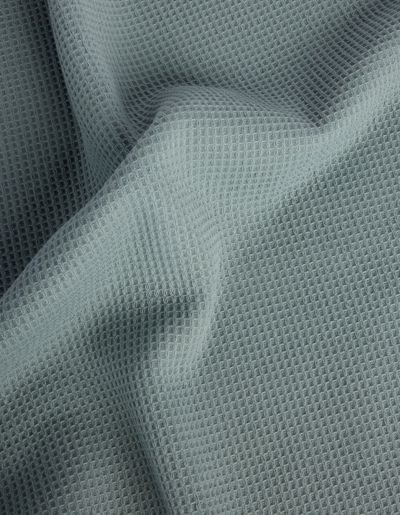 Cozy Earth’s Waffle Bath Sheets feature light gray fabric with a waffle-like texture and a grid of small, raised squares. The soft material drapes gently, creating subtle folds and shadows on the surface. 