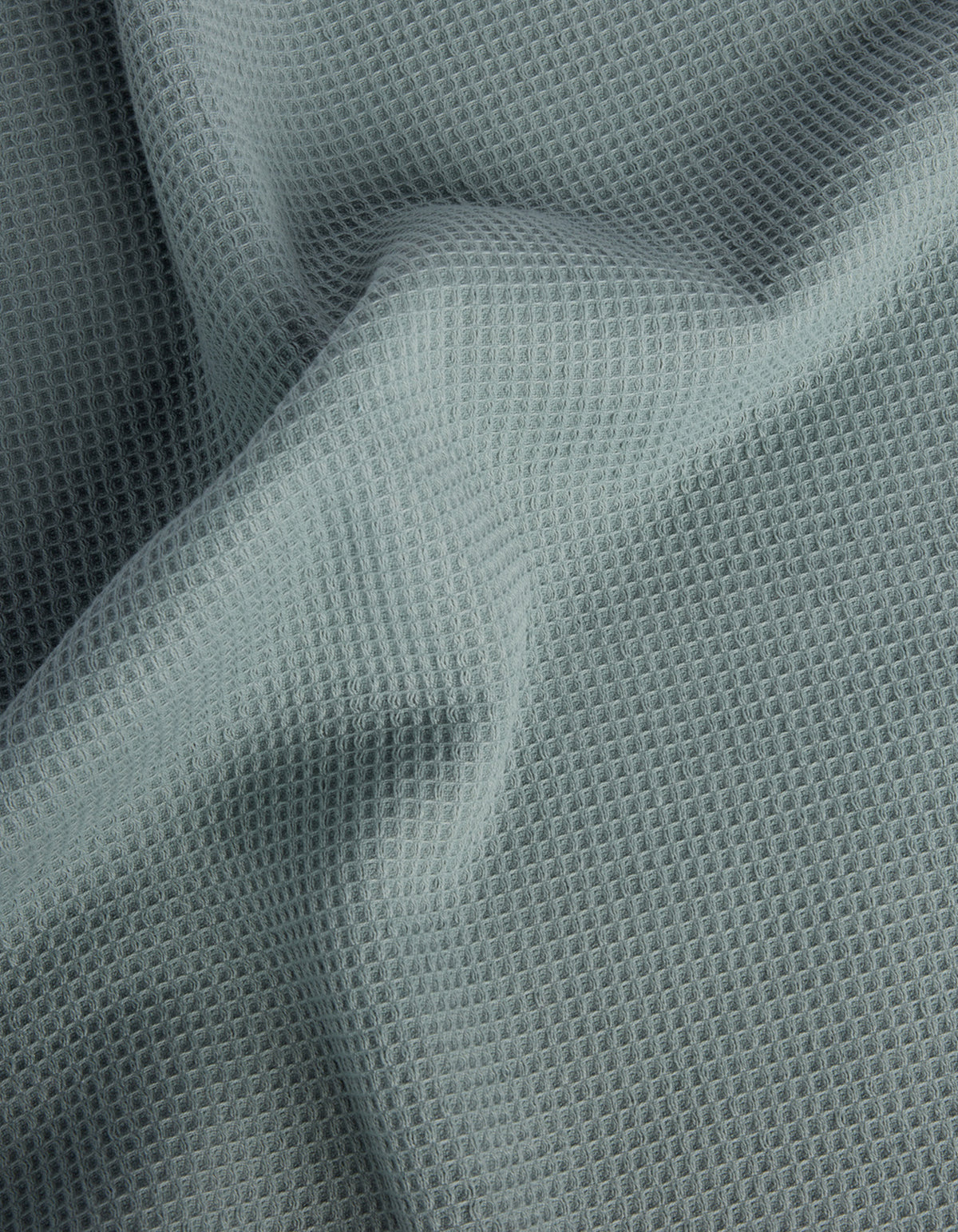 Cozy Earth’s Waffle Bath Sheets feature light gray fabric with a waffle-like texture and a grid of small, raised squares. The soft material drapes gently, creating subtle folds and shadows on the surface. |Color:Brook