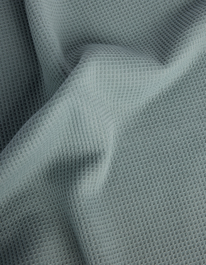 Cozy Earth’s Waffle Bath Sheets feature light gray fabric with a waffle-like texture and a grid of small, raised squares. The soft material drapes gently, creating subtle folds and shadows on the surface.