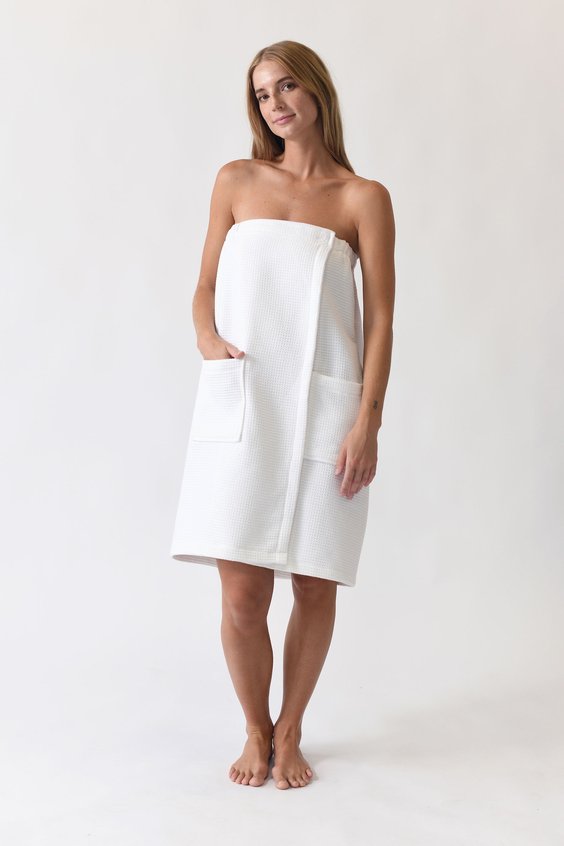 A person stands barefoot against a simple background, wearing the Cozy Earth Waffle Bath Wrap. The wrap features pockets and is secured around the chest. The person has long hair and appears relaxed. |Color:White