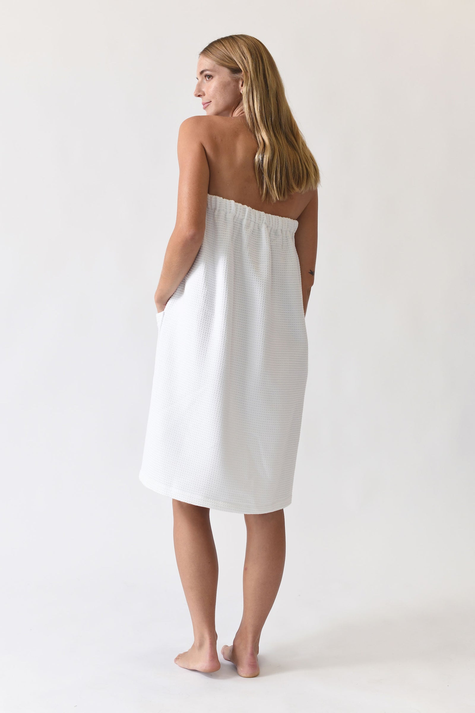 A woman with long blonde hair stands barefoot, gazing to the side while wrapped in the Cozy Earth At-Home Spa Bundle's white textured towel that covers her chest and falls to her knees, set against a plain, light-colored background.