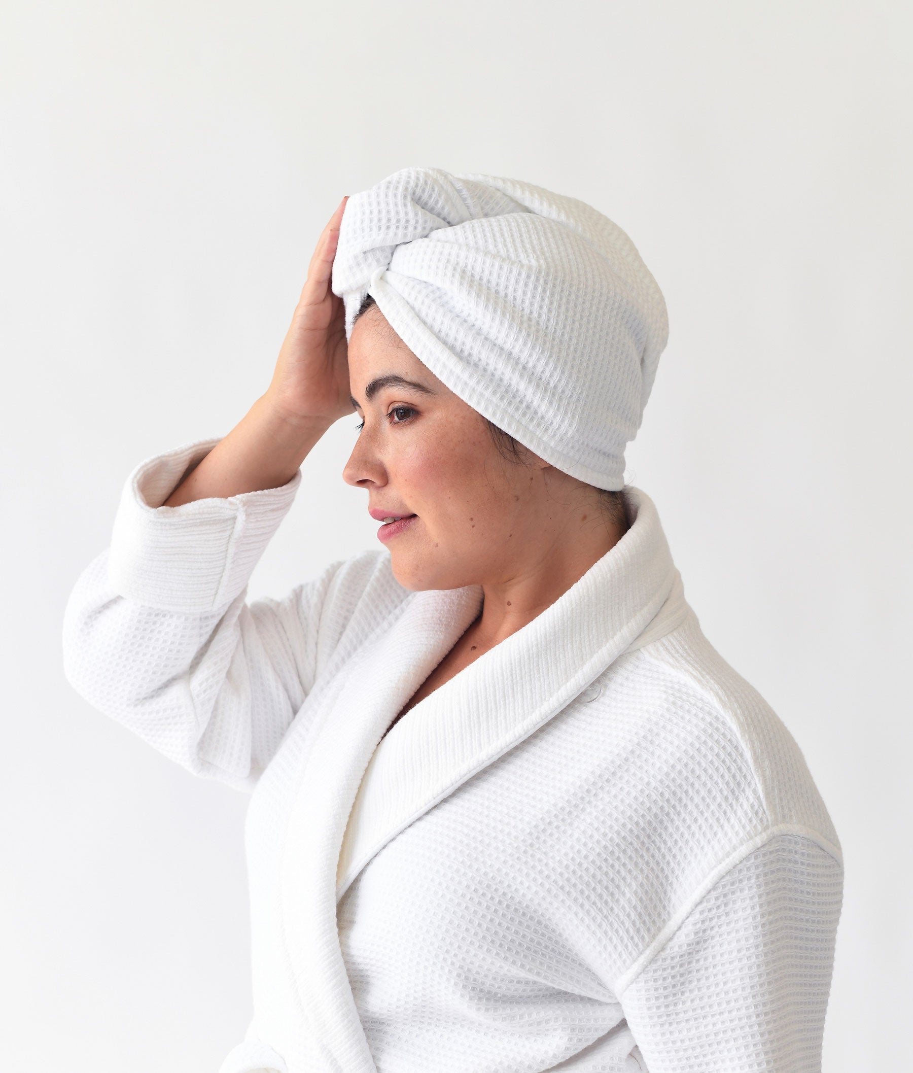 A person is wearing a Cozy Earth white bathrobe and a Waffle Hair Towel wrapped on their head, standing in profile against a plain white background, with one hand touching the towel.