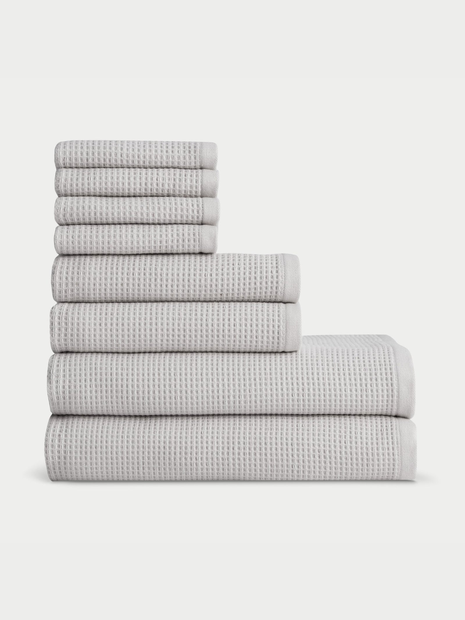 A neatly arranged set of eight folded towels from the Waffle Bath Towel Set by Cozy Earth is displayed in a pyramid formation, with the largest towels at the bottom and the smallest at the top, against a plain white background. These grey towels feature a textured, waffle-like pattern. 