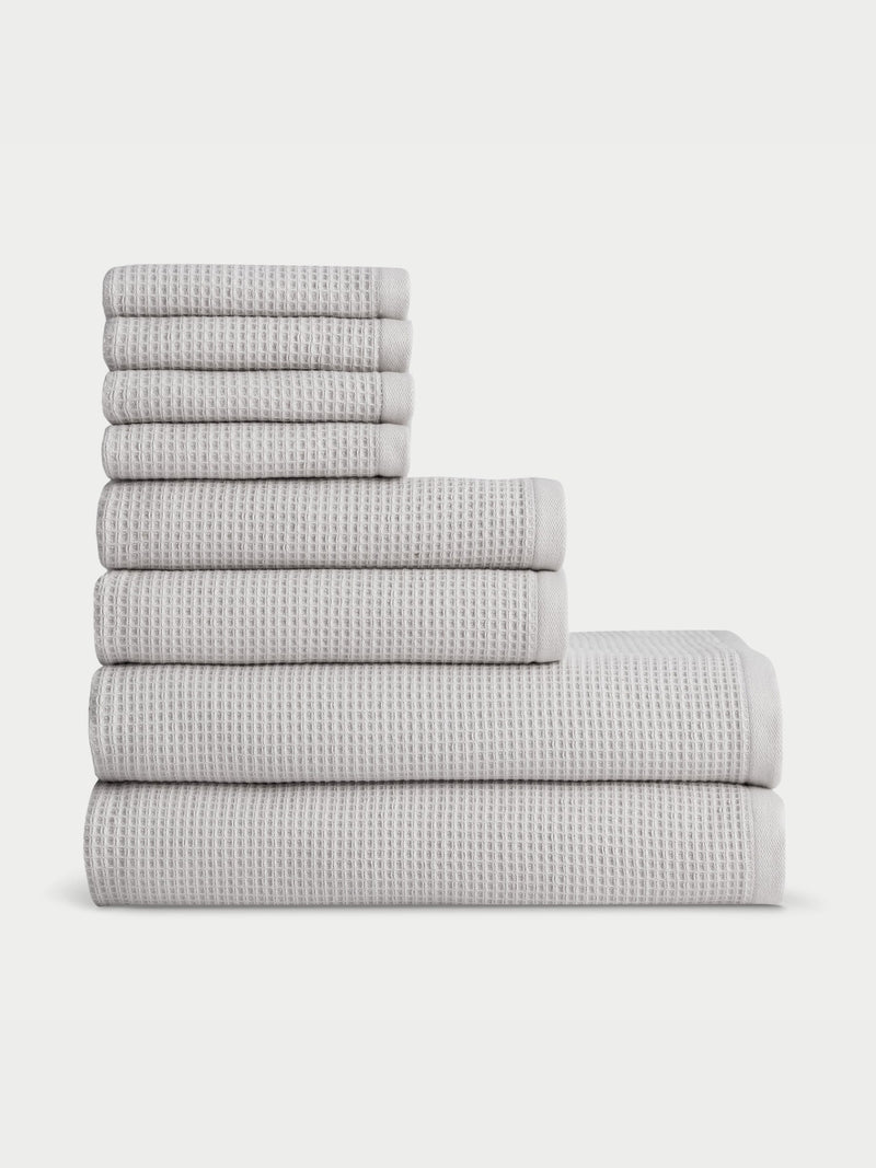 A neatly arranged set of eight folded towels from the Waffle Bath Towel Set by Cozy Earth is displayed in a pyramid formation, with the largest towels at the bottom and the smallest at the top, against a plain white background. These grey towels feature a textured, waffle-like pattern.