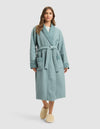 A woman with long brown hair stands facing forward in a light blue Cozy Earth Waffle Bath Robe with a belt and two front pockets, paired with beige slippers. The background is a plain light gray. |Color:Brook