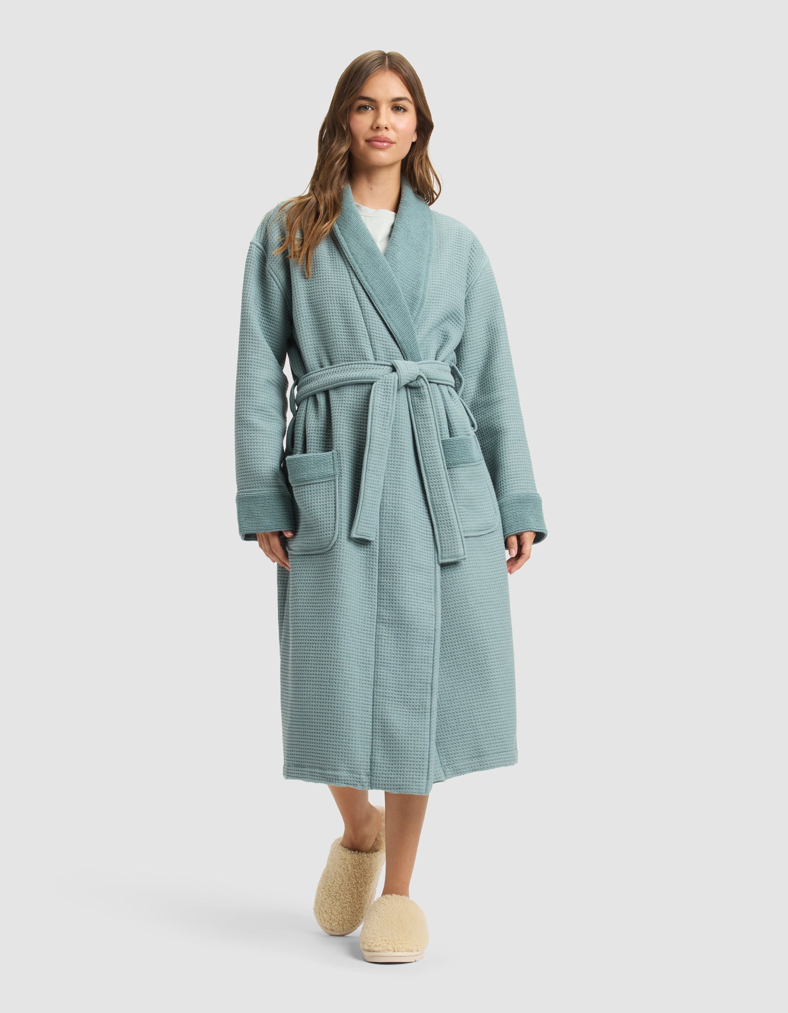 A woman with long brown hair stands facing forward in a light blue Cozy Earth Waffle Bath Robe with a belt and two front pockets, paired with beige slippers. The background is a plain light gray. |Color:Brook