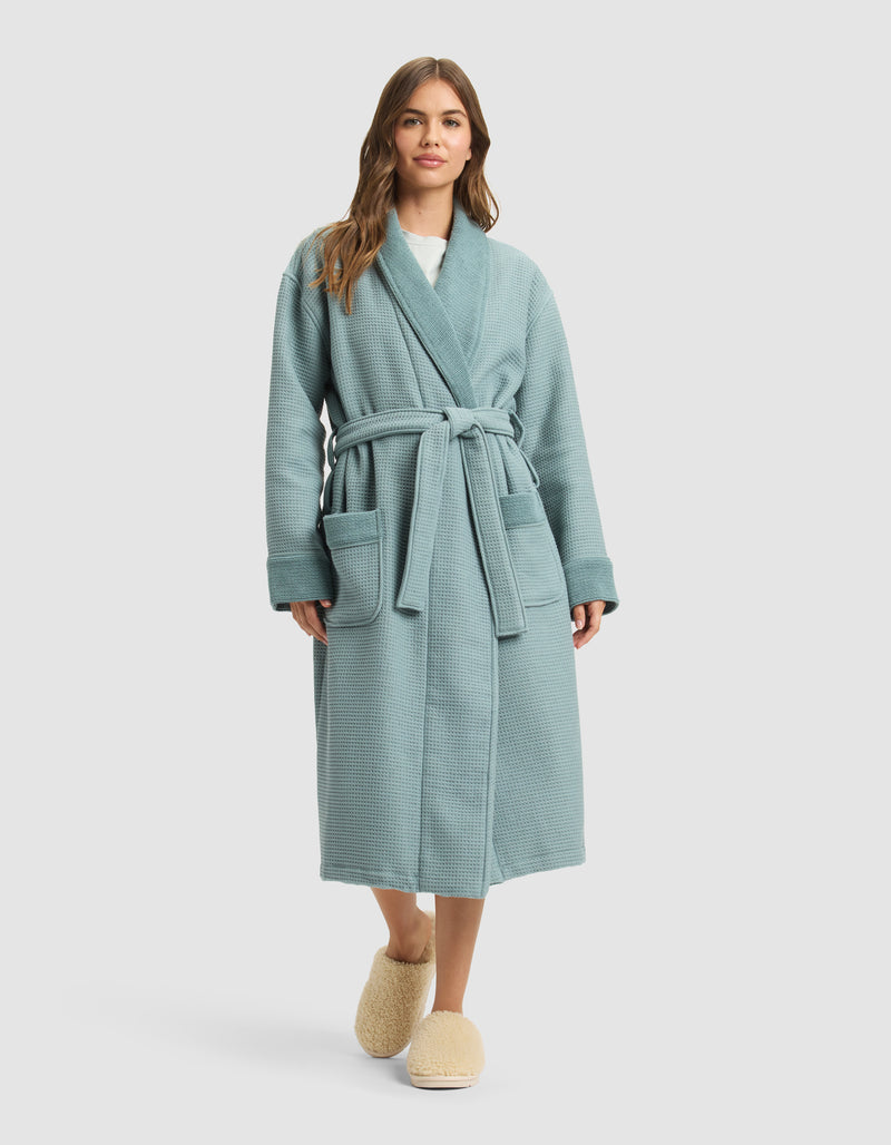A woman with long brown hair stands facing forward in a light blue Cozy Earth Waffle Bath Robe with a belt and two front pockets, paired with beige slippers. The background is a plain light gray.