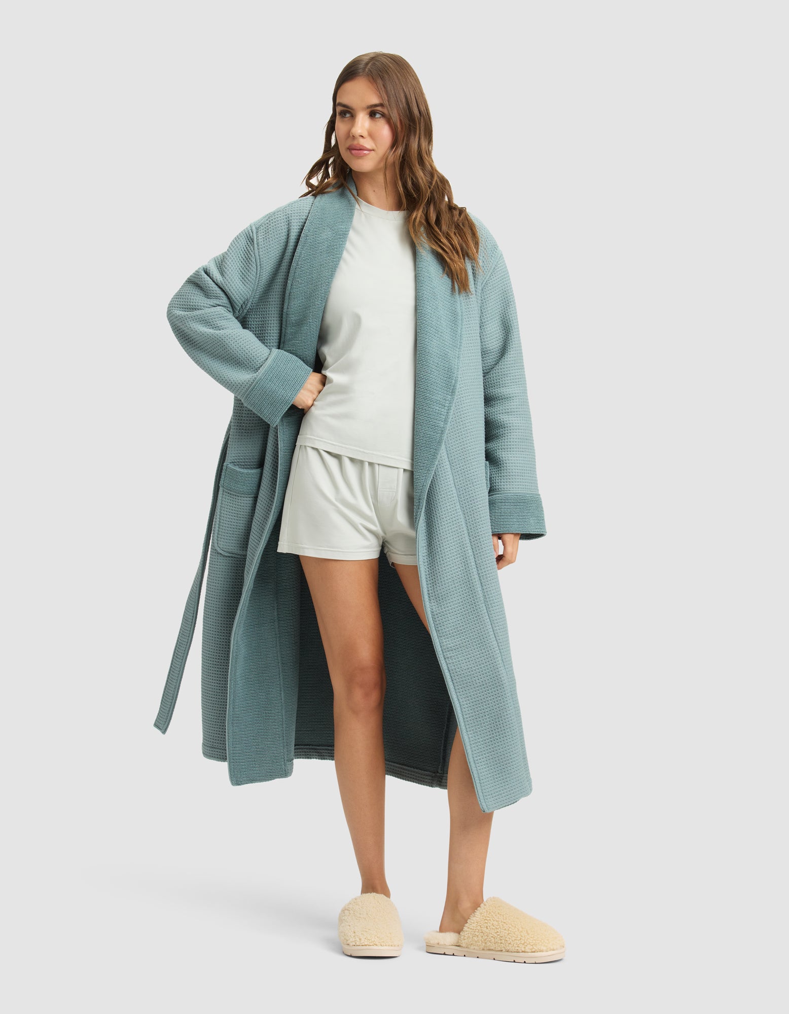 A woman with wavy brown hair models the Cozy Earth Waffle Bath Robe in teal over a white T-shirt and shorts with beige slippers, posing against a plain light gray background. 