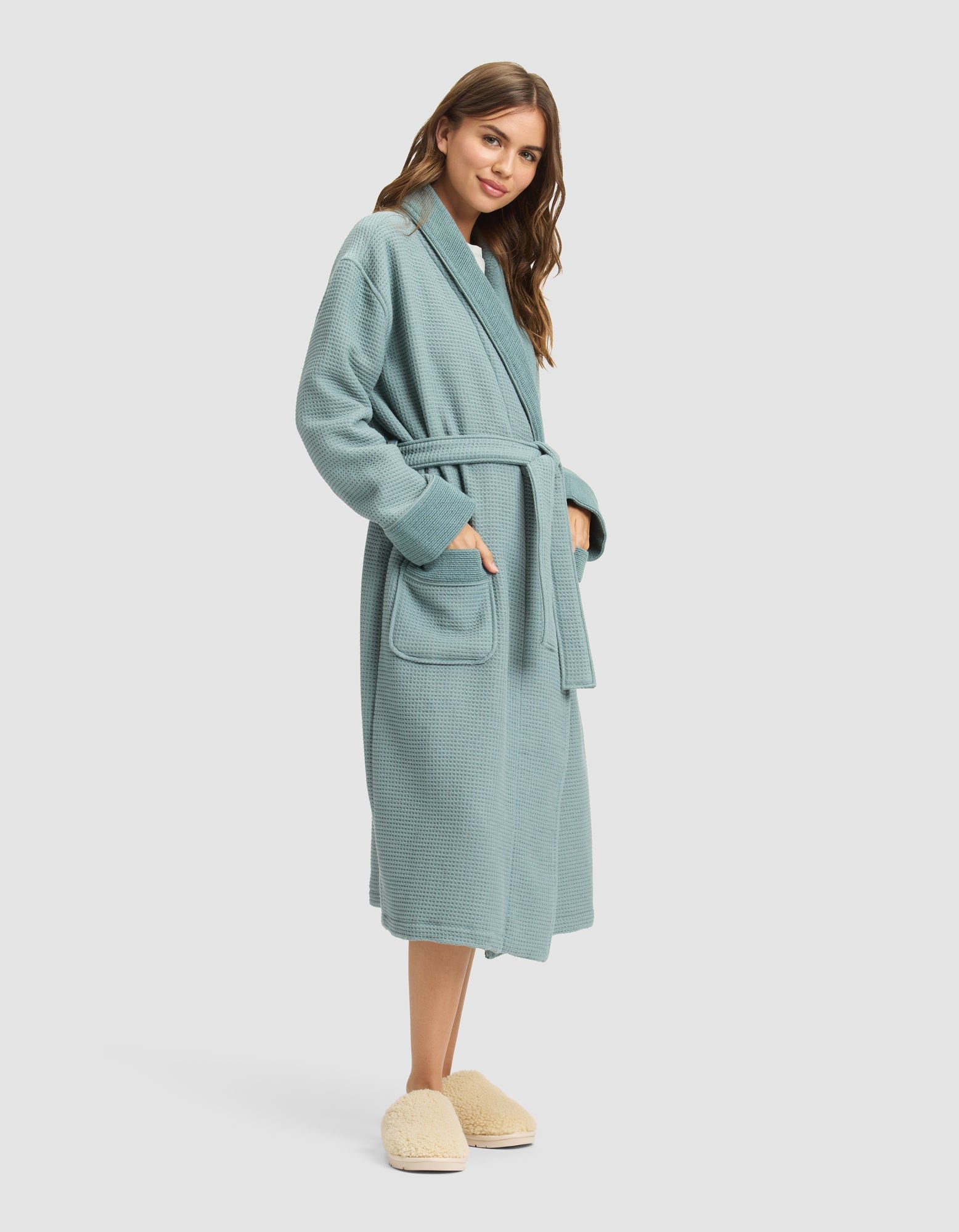 A woman with long brown hair, smiling slightly and facing right, wears a Cozy Earth Waffle Bath Robe in light blue with a belt and large front pockets, paired with fuzzy beige slippers. 