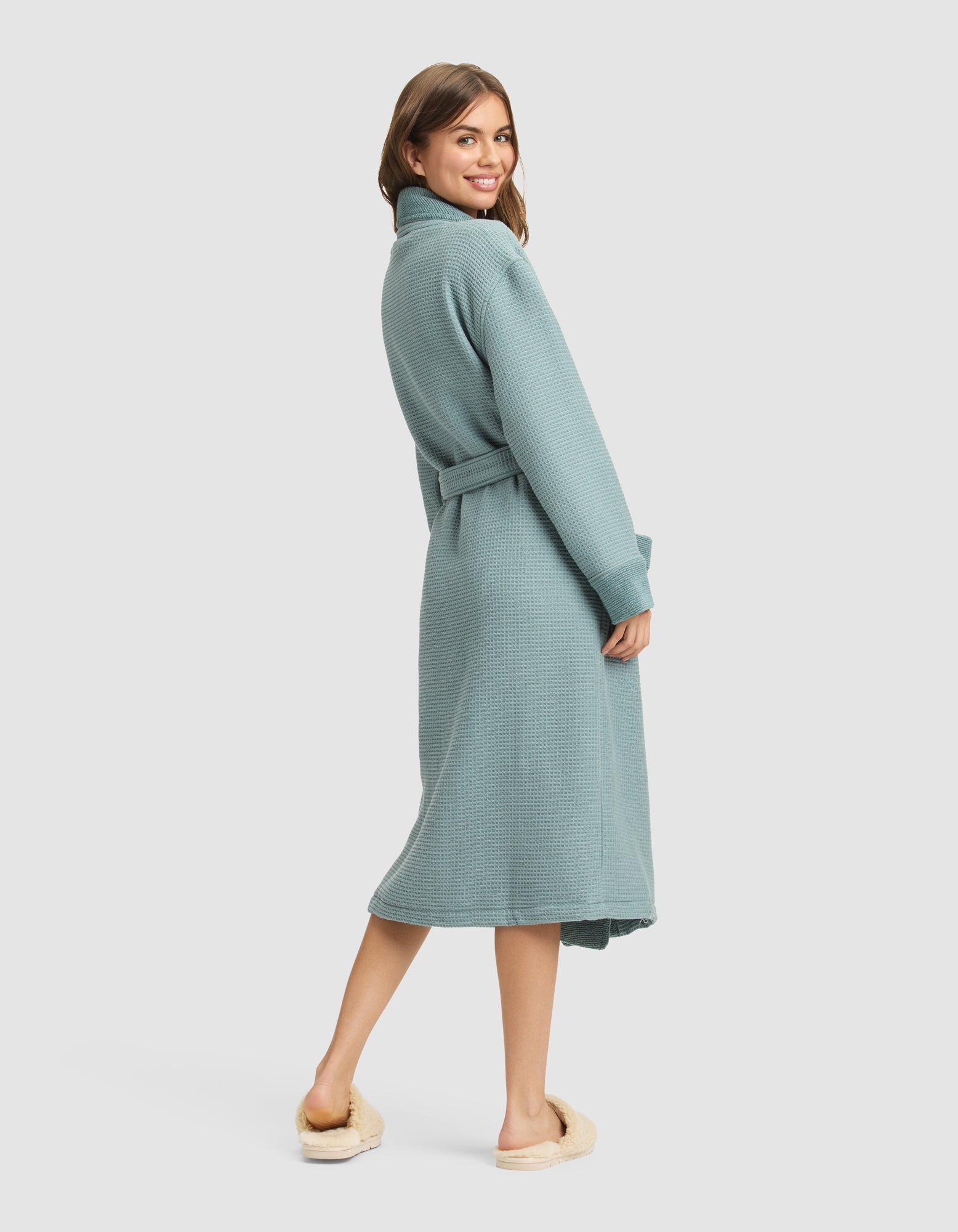 A woman smiles over her shoulder, wearing the Cozy Earth Waffle Bath Robe in light blue, tied at the waist, with cream slippers, standing against a plain light background. 