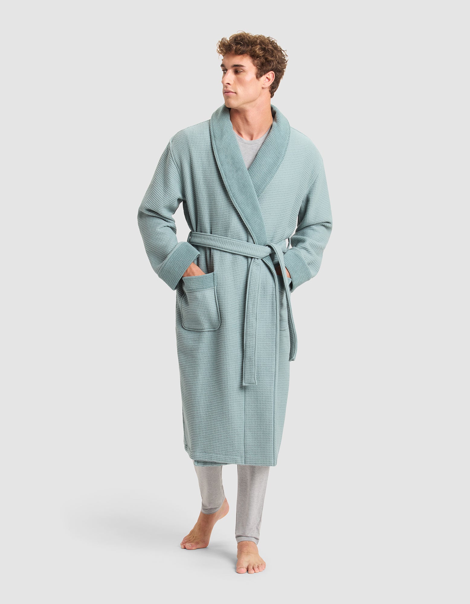 A man stands barefoot, looking to the side, wearing Cozy Earth’s Waffle Bath Robe in light blue with a belt and two front pockets over light gray pants.  