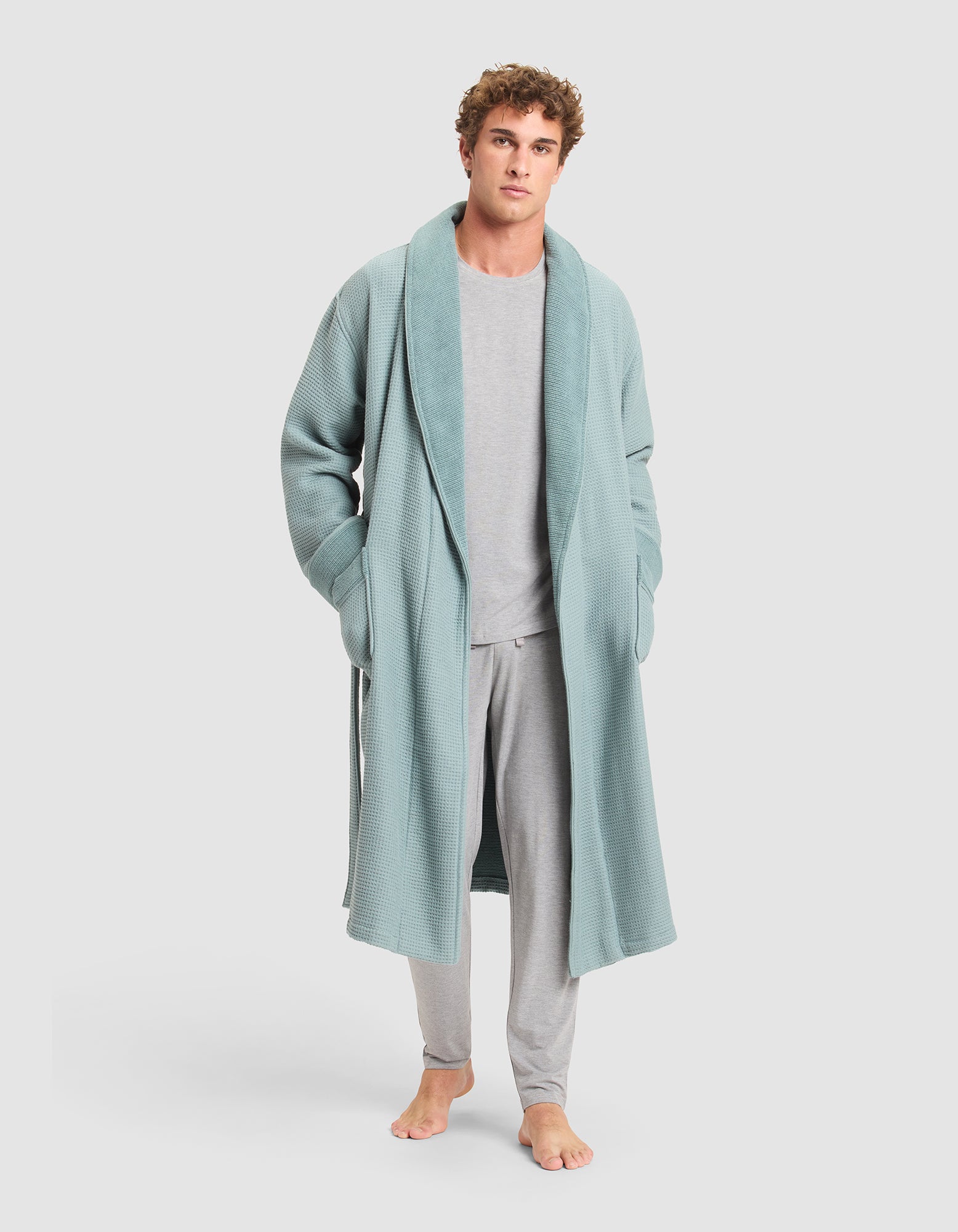 A man stands barefoot in light gray pajamas and a Cozy Earth Waffle Bath Robe with pockets, looking at the camera against a plain white background.  