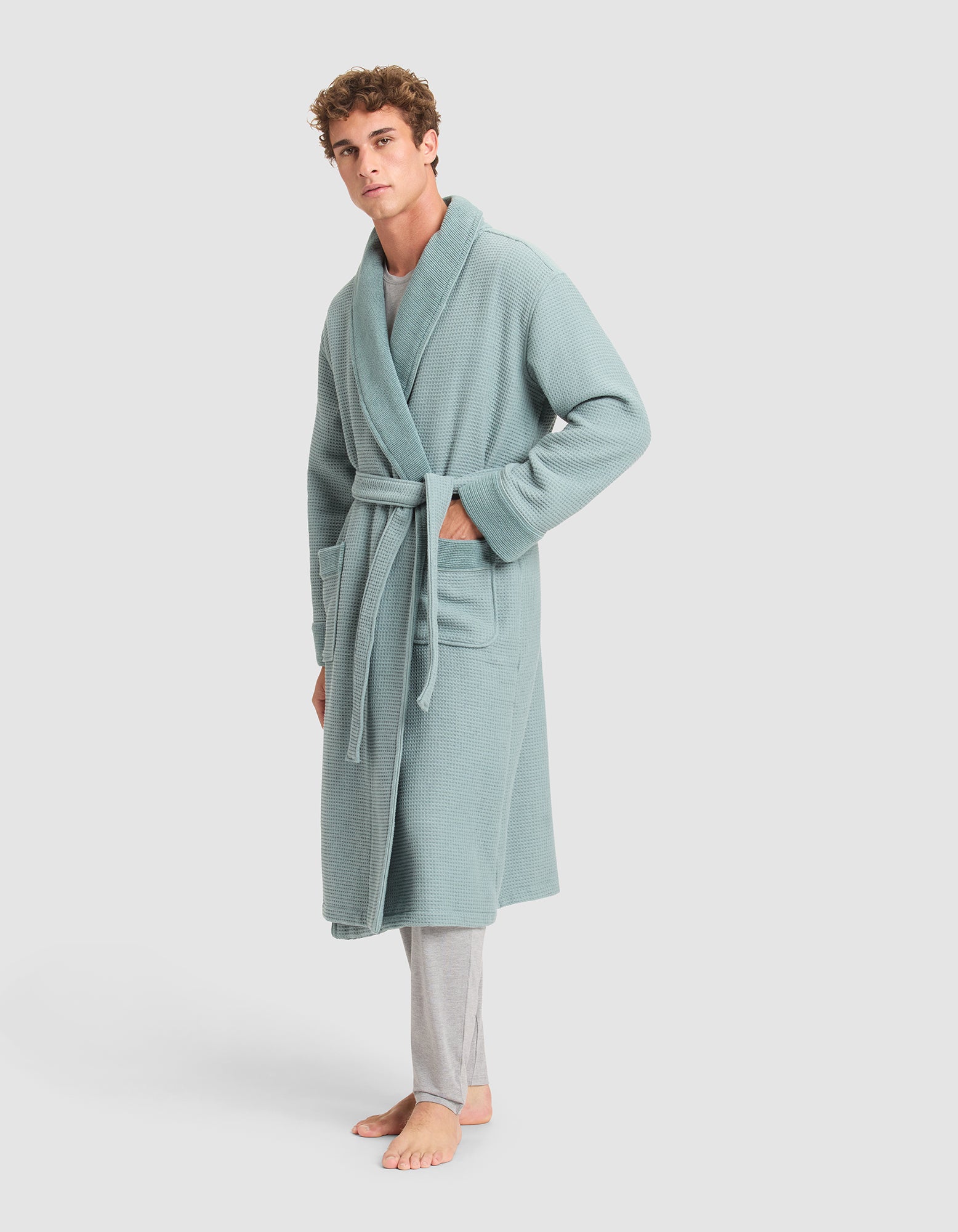 A man stands barefoot in Cozy Earth's Waffle Bath Robe, a light blue-green, waffle-knit robe with a belt and two front pockets, paired with light gray pajama pants, against a plain light background.  
