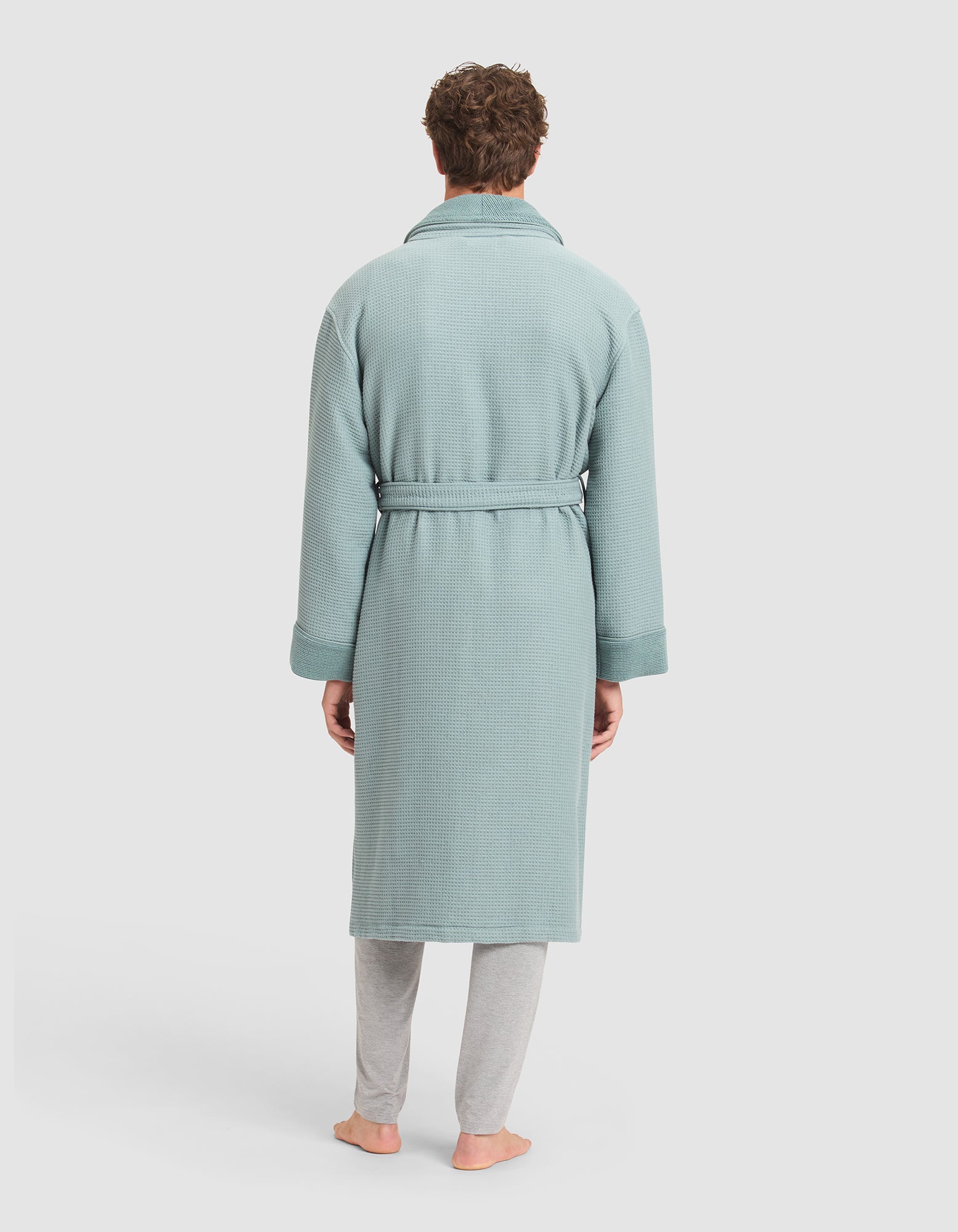 A person stands barefoot facing away, wearing the Cozy Earth Waffle Bath Robe with a belt over light gray pants, against a plain white background.  
