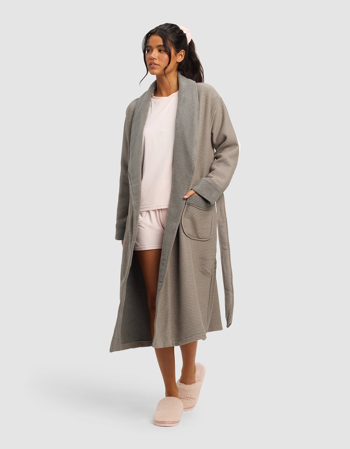 A woman stands against a plain background, wearing the Cozy Earth Waffle Bath Robe in gray over a light pink pajama set and pink fuzzy slippers, with one hand in her pocket and a relaxed expression.