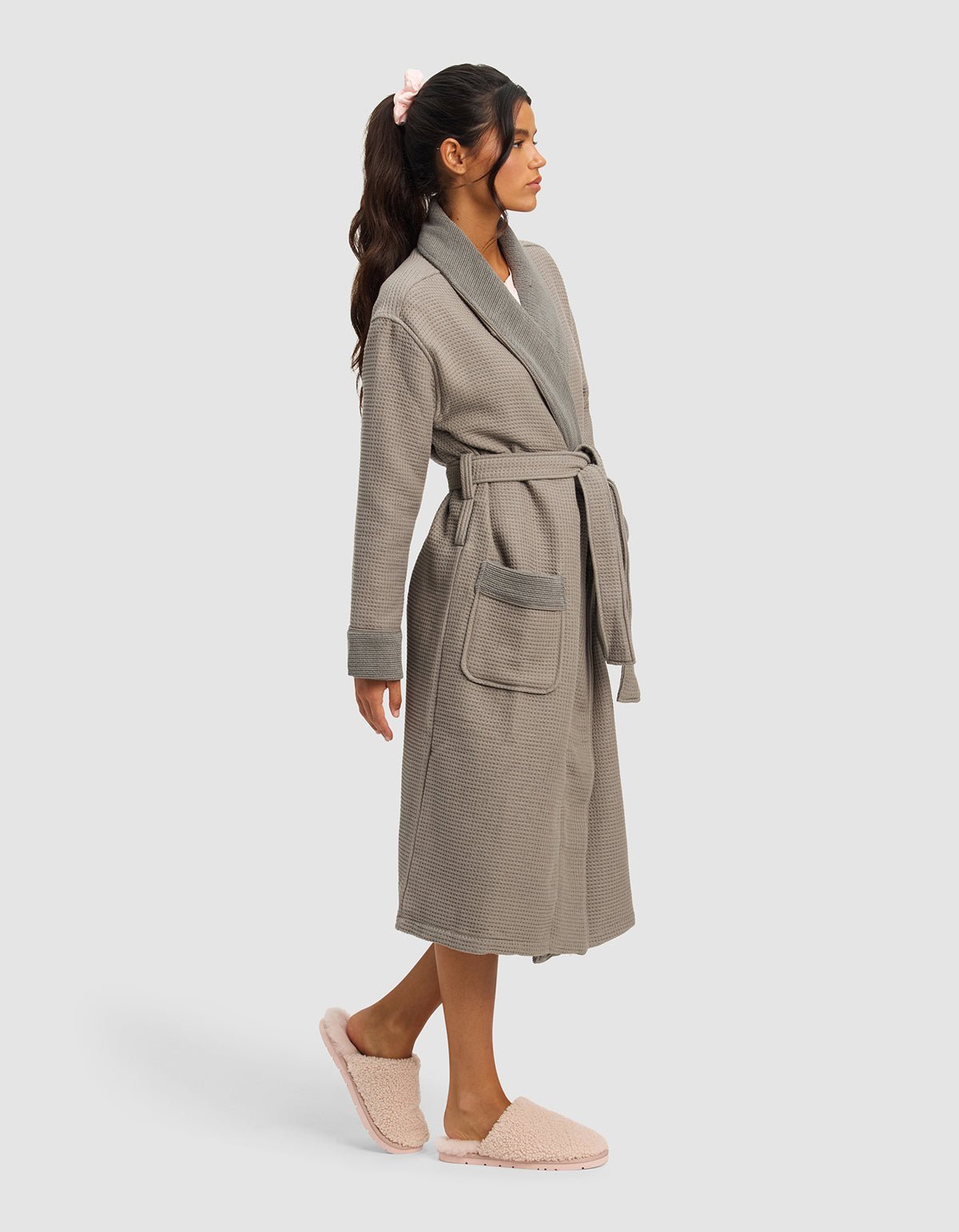 A woman with long dark hair in a ponytail wears the Cozy Earth Waffle Bath Robe in light grey, featuring large pockets and a belt, along with pink fuzzy slippers. She stands in profile against a plain light background.|Color:Charcoal