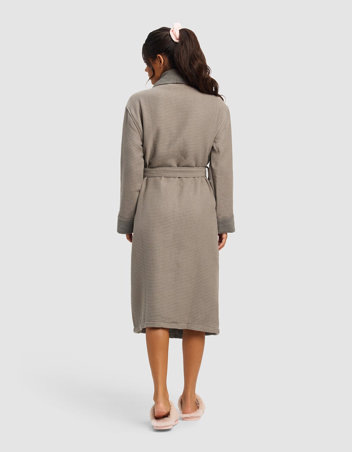 A woman faces away, wearing the Cozy Earth Waffle Bath Robe in textured gray with a belt and fuzzy pink slippers. Her long dark hair is tied back with a light pink scrunchie against a plain light background.