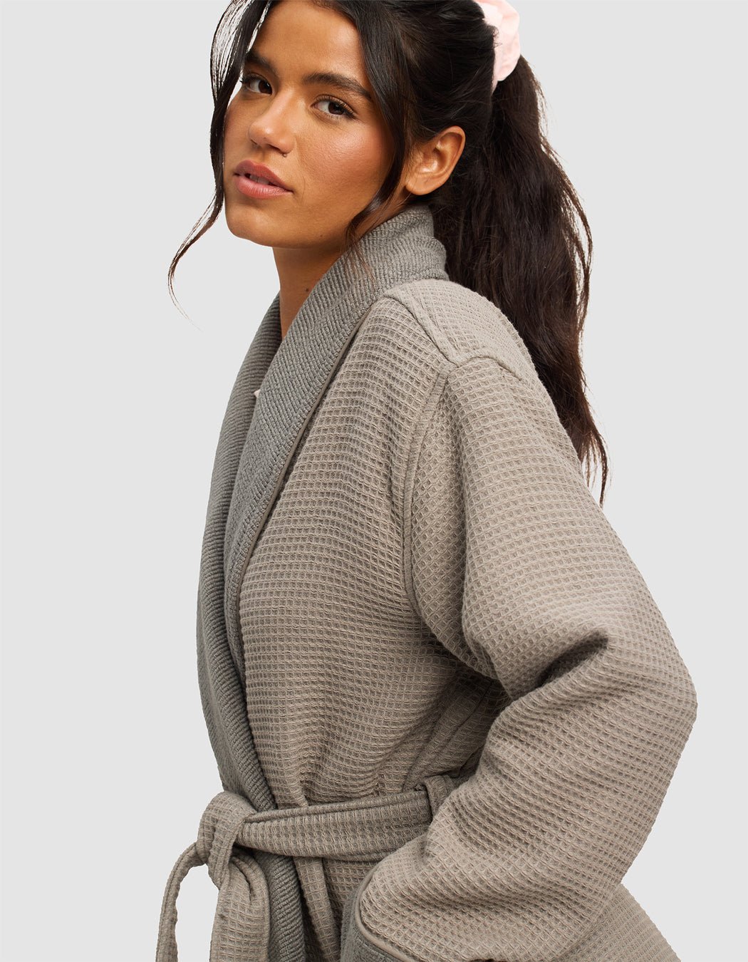 A woman with long dark hair in a ponytail, wearing a light pink scrunchie and the Cozy Earth Waffle Bath Robe with belt, poses against a plain light gray background.