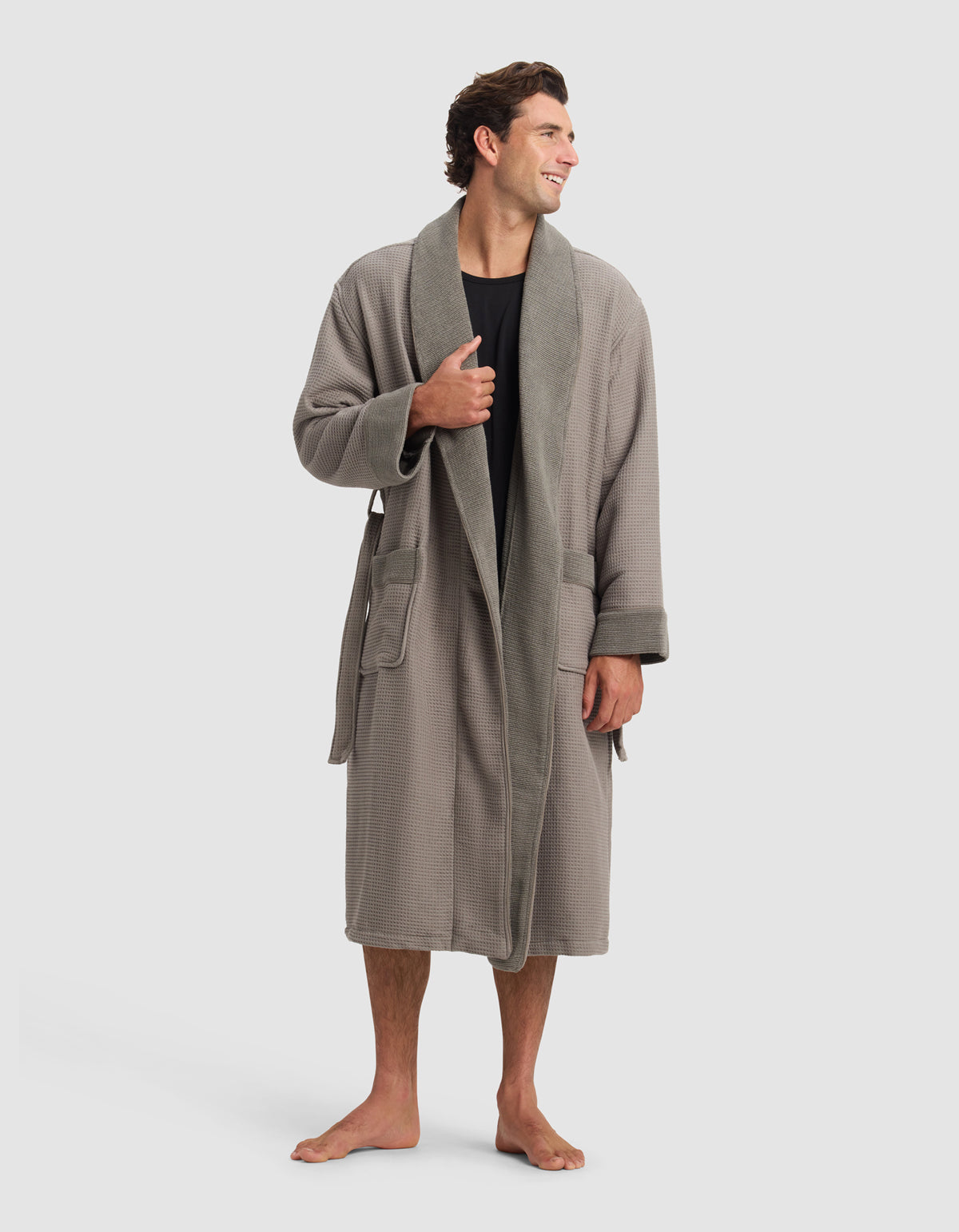A man stands barefoot, smiling and looking to the side, wearing Cozy Earth's Waffle Bath Robe in gray over a black shirt. The robe features large pockets, a belt, and is set against a plain light gray background.