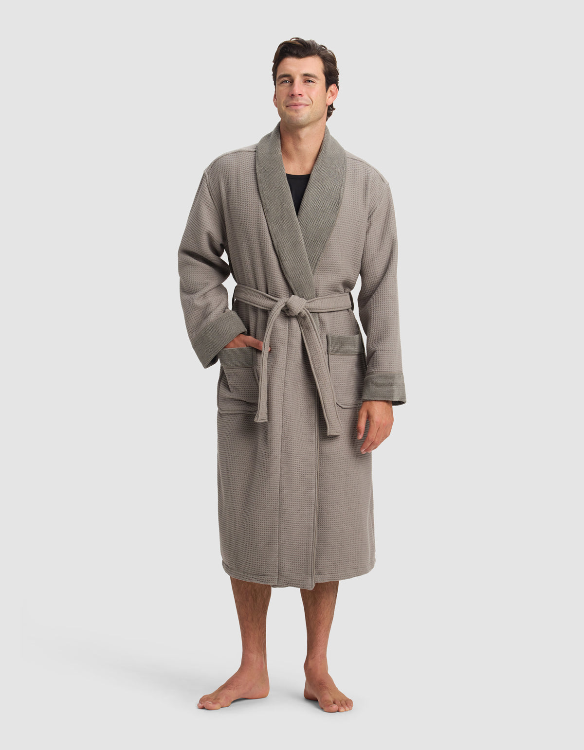 A man stands barefoot in a Cozy Earth Waffle Bath Robe—light brown with a darker shawl collar, front pockets, and a tied belt—worn over a black shirt. The background is plain and light-colored.
