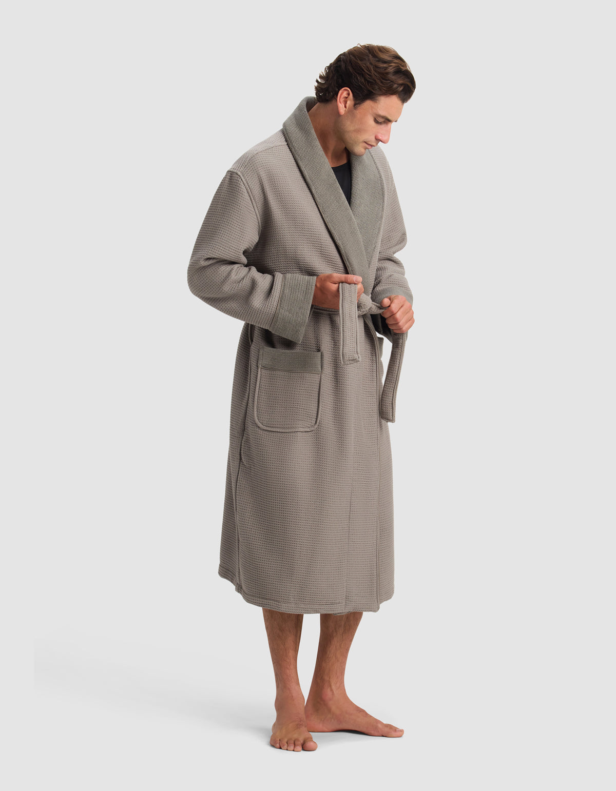 A man barefoot in a Cozy Earth Waffle Bath Robe stands against a plain light background, looking down as he ties the robe's belt at his waist.