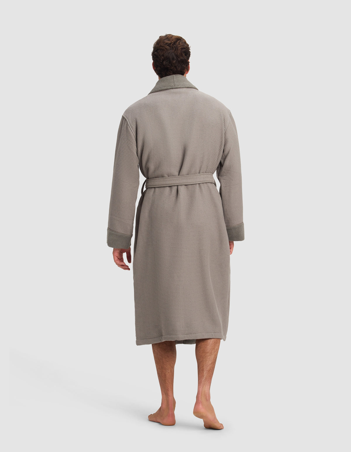 A man stands barefoot, facing away from the camera, wearing the Cozy Earth Waffle Bath Robe—a long, light gray, textured robe with a belted waist.