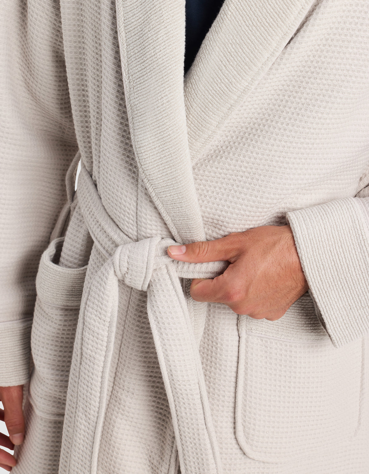 A person is shown tying the belt of a Cozy Earth Waffle Bath Robe at the waist. The light-colored, textured robe features large front pockets, with only the wearer’s torso and hands visible.