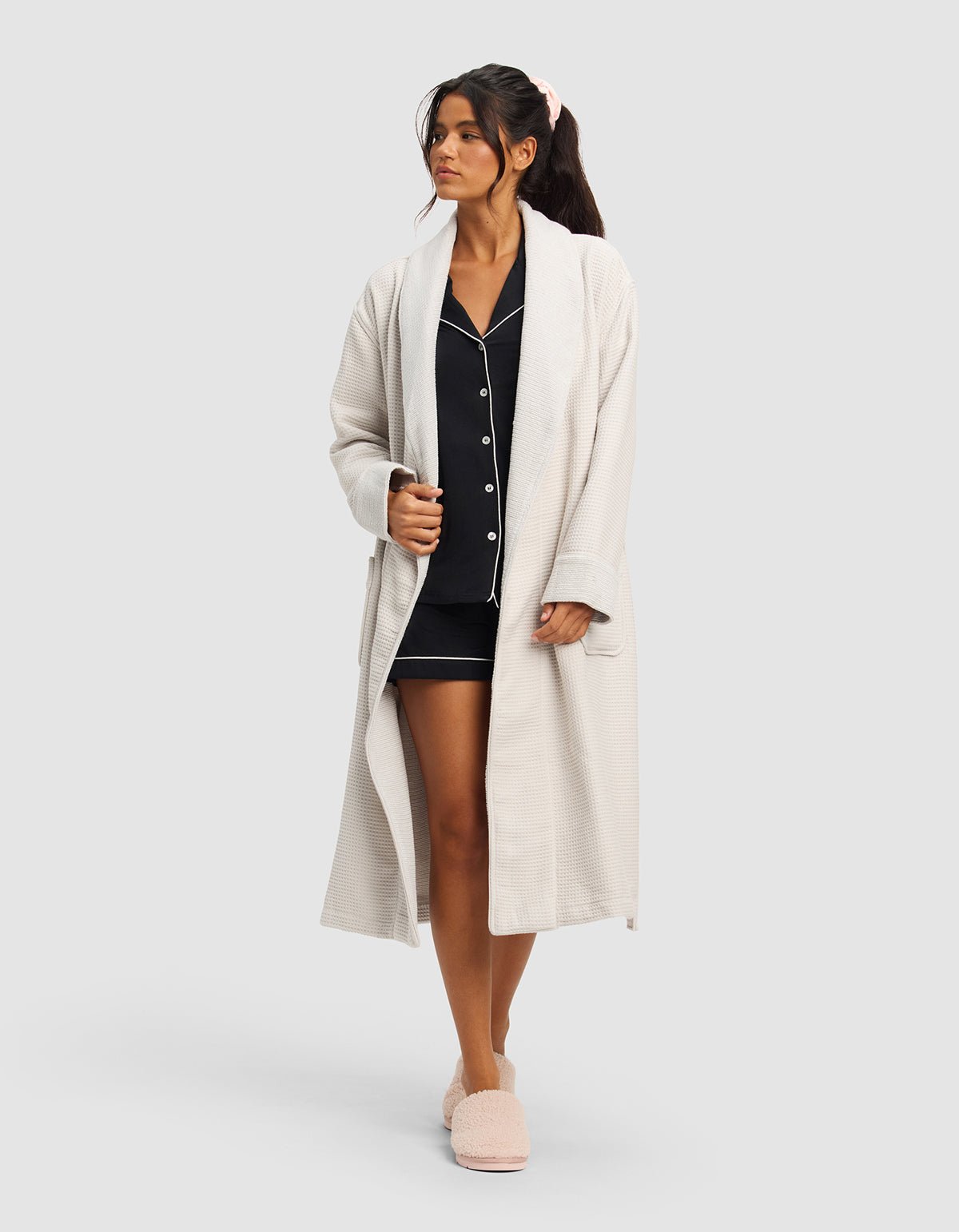 A woman wears the Cozy Earth Waffle Bath Robe over dark pajamas with white trim and matching slippers, her hair tied with a pink scrunchie as she gazes slightly to the side against a plain background.|Color:Light Grey