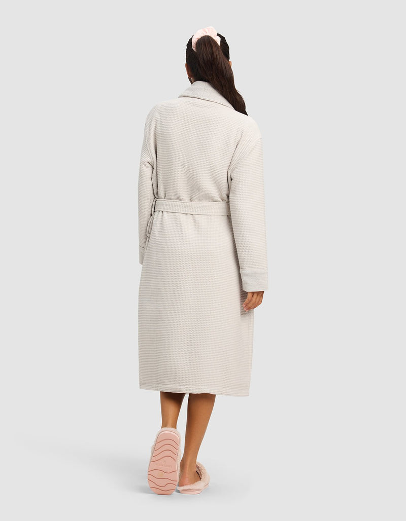 A woman seen from behind wears the Cozy Earth Waffle Bath Robe in light beige with a belt, pink slippers, and a light pink scrunchie, standing against a plain light gray background.