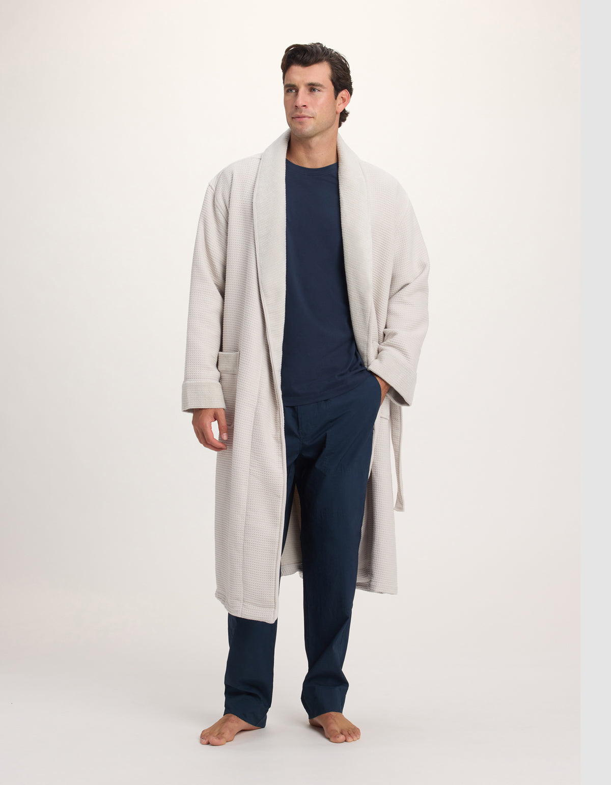 A man stands barefoot on a light background wearing the Cozy Earth Waffle Bath Robe in cream over a navy blue shirt and pants, looking slightly to the side with his hands relaxed by his sides.